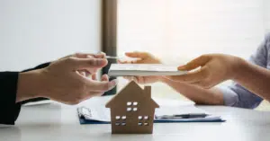 One person handing over money and documents to another, with a house in the background, symbolizing the process of paying the deposit for a property purchase.