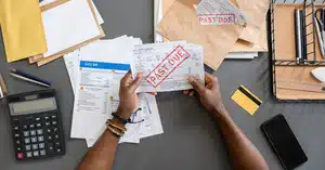 Close-up of hands holding a 'PAST DUE' bill with various envelopes, documents, a calculator, and a phone on a desk.