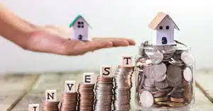 Stacks of coins spelling “INVEST” with small model houses, representing saving for an investment property deposit and building wealth.
