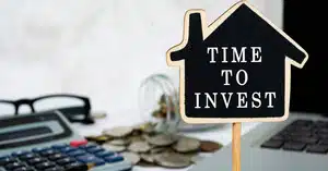 House-shaped sign reading “Time to Invest” next to coins and a calculator, symbolizing planning for an investment property deposit.