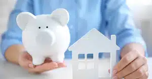 Person holding a white piggy bank and a cutout house, symbolizing saving 10% for an investment property deposit.