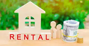 Wooden house model with “RENTAL” blocks, family figurines, and money roll, representing rental income from an investment property deposit.